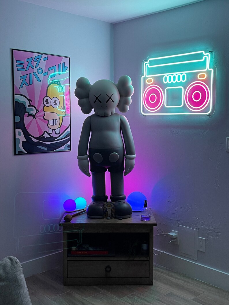 K&ouml;nnte beinhalten: Eine graue und wei&szlig;e Figur eines Comic-Charakters mit einem 'X' auf der Stirn steht auf einem Holztisch mit zwei blau und lila leuchtenden Kugeln. Ein Neon-Schild eines Boombox mit rosa und blauen Lichtern befindet sich an der Wand hinter der Figur. Ein Poster mit japanischem Text und einem Comic-Charakter befindet sich an der Wand links von der Figur.