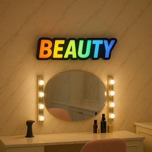 Könnte beinhalten: Ein Schminkspiegel mit einem regenbogenfarbenen "BEAUTY"-Schild darüber. Der Spiegel hat einen goldenen Rahmen und ist auf jeder Seite von fünf Glühbirnen umgeben.