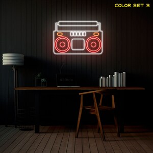 K&ouml;nnte beinhalten: Wei&szlig;er Neon-Umriss eines Boombox mit roten Neon-Akzenten. Der Boombox befindet sich an einer schwarzen Wand mit einem Holztisch und einem Stuhl davor. Der Text "COLOR SET 3" befindet sich in der oberen rechten Ecke des Bildes. Der Text "NEONIX" befindet sich auf einem Laptop auf dem Tisch.
