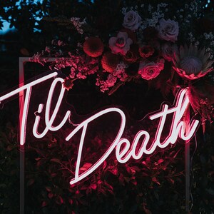 May include: A pink neon sign with the words "Til Death" in a cursive font. The sign is framed in a white square frame and is surrounded by a floral arrangement of pink and red flowers.
