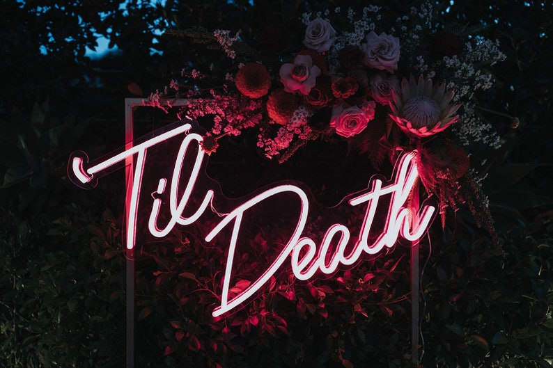 May include: A pink neon sign with the words "Til Death" in a cursive font. The sign is framed in a white square frame and is surrounded by a floral arrangement of pink and red flowers.
