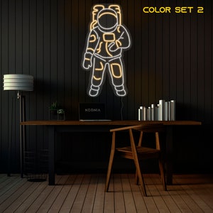 May include: A white neon astronaut outline with a yellow glow, mounted on a black wall. The neon sign is in a room with a desk, chair, and lamp. The text "COLOR SET 2" is in the upper right corner of the image.