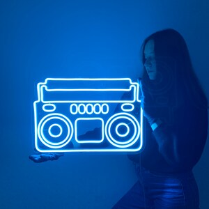 K&ouml;nnte beinhalten: Eine Person h&auml;lt ein blaues Neon-Schild in Form einer alten Boombox. Das Schild leuchtet und die Person steht vor einer blauen Wand.