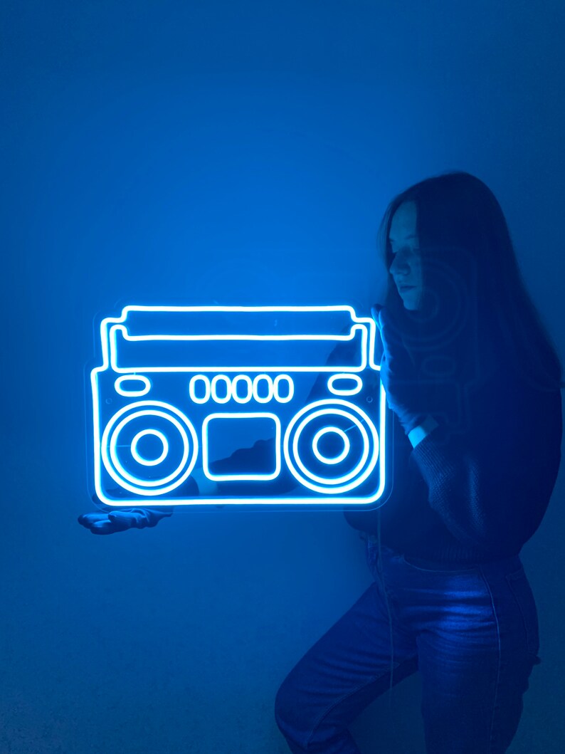 K&ouml;nnte beinhalten: Eine Person h&auml;lt ein blaues Neon-Schild in Form einer alten Boombox. Das Schild leuchtet und die Person steht vor einer blauen Wand.