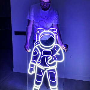 May include: A white neon astronaut sign with a blue glow. The sign is mounted on a clear acrylic panel and is being held by a person.