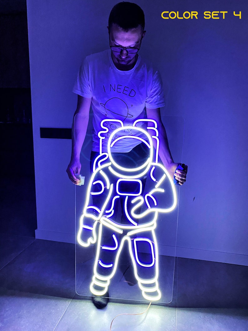 May include: A white neon astronaut sign with a blue glow. The sign is mounted on a clear acrylic panel and is being held by a person.