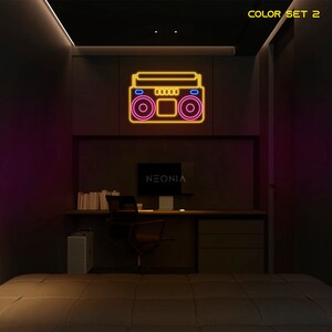 K&ouml;nnte beinhalten: Ein Neon-Schild in Form eines Boombox mit gelben und rosa Umrissen. Das Schild ist an einer schwarzen Wand in einem Raum mit einem Schreibtisch und einem Bett angebracht. Das Schild lautet "NEONIA" in der Mitte unten.