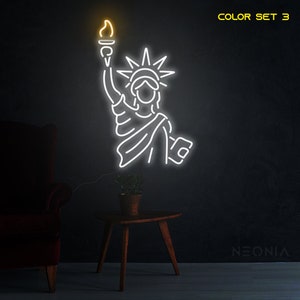 May include: White neon sign depicting the Statue of Liberty with a torch in her raised right hand. The sign is lit up and has a yellow glow. The text "COLOR SET 3" is in the top right corner of the image. The text "NEONIA" is in the bottom right corner of the image.
