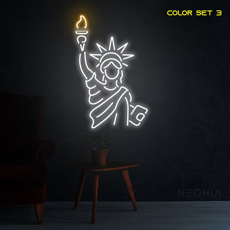 May include: White neon sign depicting the Statue of Liberty with a torch in her raised right hand. The sign is lit up and has a yellow glow. The text "COLOR SET 3" is in the top right corner of the image. The text "NEONIA" is in the bottom right corner of the image.