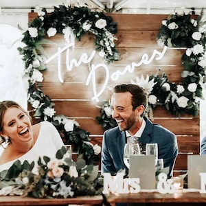 May include: A wedding reception with a wooden backdrop decorated with greenery and white flowers. The couple is seated at a table with white signs that read "Mrs." and "Mr." A neon sign with the text "The Domes" is hanging on the wall.