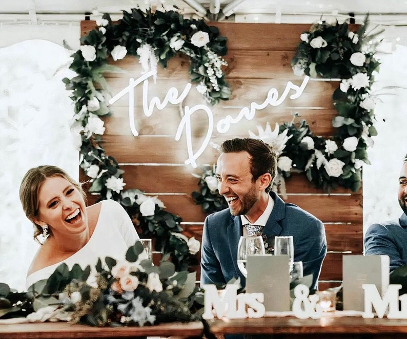 May include: A wedding reception with a wooden backdrop decorated with greenery and white flowers. The couple is seated at a table with white signs that read "Mrs." and "Mr." A neon sign with the text "The Domes" is hanging on the wall.