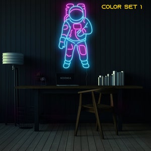 May include: Neon sign of an astronaut in a blue and pink outline against a dark wood background. The sign says "NEONIA" at the bottom. The text "COLOR SET 1" is in the top right corner of the image.