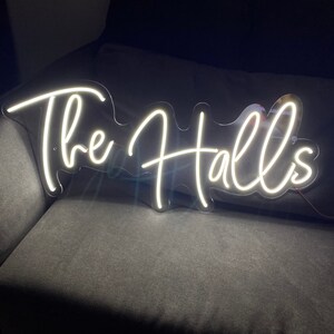 May include: White neon sign with the text "The Halls" on a clear acrylic background. The sign is sitting on a dark gray couch.