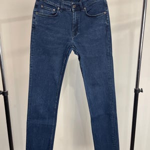 May include: Dark blue denim jeans with a straight leg cut and a raw hem. The jeans have a classic five-pocket design and a button and zipper closure. The brand name "Acne Studios" is visible on the label.