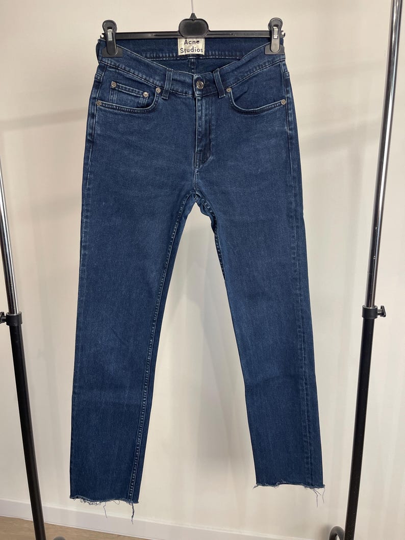 May include: Dark blue denim jeans with a straight leg cut and a raw hem. The jeans have a classic five-pocket design and a button and zipper closure. The brand name "Acne Studios" is visible on the label.