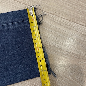 May include: A section of dark blue denim fabric with a frayed edge is measured with a yellow tape measure. The tape measure shows a measurement of approximately 6 inches.