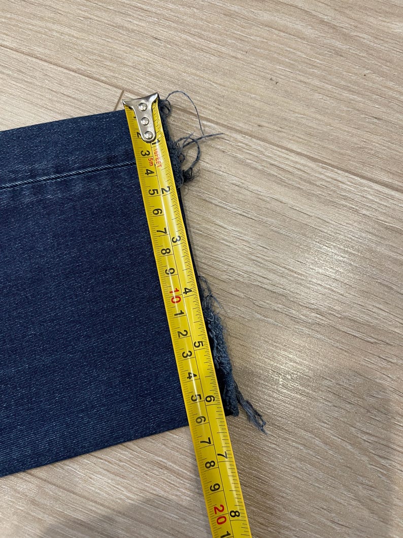 May include: A section of dark blue denim fabric with a frayed edge is measured with a yellow tape measure. The tape measure shows a measurement of approximately 6 inches.