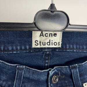 May include: Close-up of dark blue denim jeans with a button closure and metal rivets. The jeans are hanging on a black hanger, with a white tag that reads "Acne Studios". The denim has a classic, worn look.