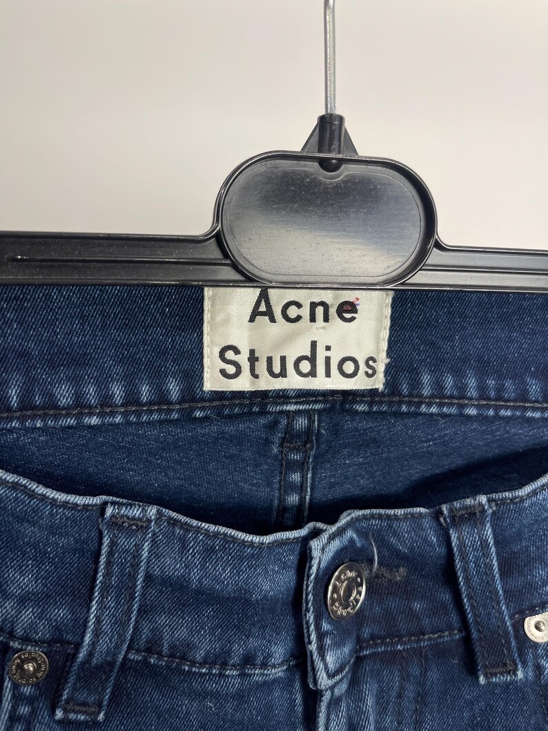 May include: Close-up of dark blue denim jeans with a button closure and metal rivets. The jeans are hanging on a black hanger, with a white tag that reads "Acne Studios". The denim has a classic, worn look.