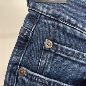 May include: Close-up of dark blue denim jeans. The image highlights the pocket details, including the stitching and metal rivets. The jeans are folded over a hanger.