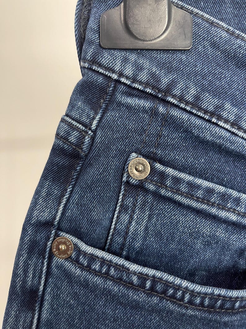 May include: Close-up of dark blue denim jeans. The image highlights the pocket details, including the stitching and metal rivets. The jeans are folded over a hanger.