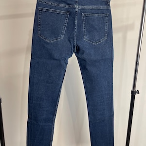 May include: Dark blue denim jeans with a classic five-pocket design. The jeans have a straight leg cut and a raw, frayed hem. The jeans are hanging on a black hanger.