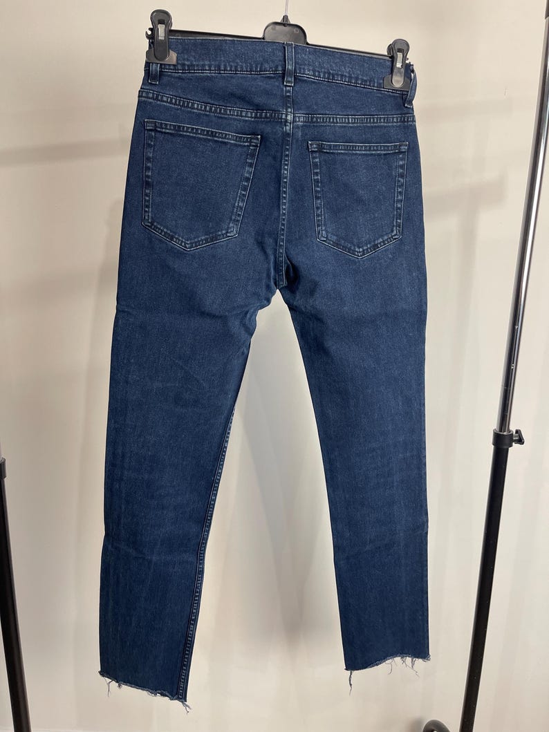 May include: Dark blue denim jeans with a classic five-pocket design. The jeans have a straight leg cut and a raw, frayed hem. The jeans are hanging on a black hanger.