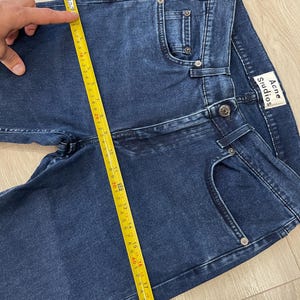 May include: Dark blue denim jeans with a classic five-pocket design. The jeans are being measured with a yellow measuring tape. The brand label "Acne Studios" is visible on the back pocket. The jeans appear to be new.
