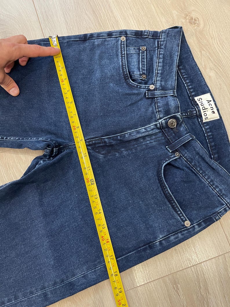 May include: Dark blue denim jeans with a classic five-pocket design. The jeans are being measured with a yellow measuring tape. The brand label "Acne Studios" is visible on the back pocket. The jeans appear to be new.