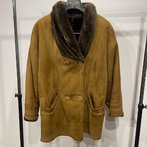 May include: A tan suede coat with a dark green fur collar and lining. The coat features a double-breasted design with large wooden buttons, two front pockets, and a classic, warm style. The coat is hanging on a metal clothing rack.