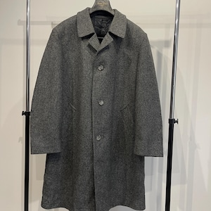 May include: A gray wool coat with a classic design hangs on a metal clothing rack. The coat features a notched collar, three buttons, and two front pockets. The coat is a medium length and appears to be made of a textured wool fabric.