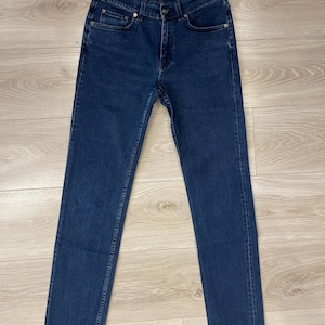 May include: Dark blue denim jeans with a straight leg and a raw hem. The jeans have a button and zipper closure, classic five-pocket design, and the "Acne Studios" label. The jeans are laid flat on a light wood surface.
