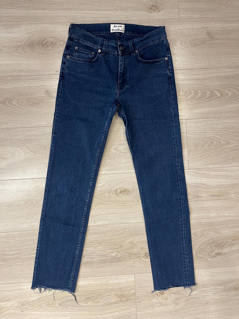 May include: Dark blue denim jeans with a straight leg and a raw hem. The jeans have a button and zipper closure, classic five-pocket design, and the "Acne Studios" label. The jeans are laid flat on a light wood surface.