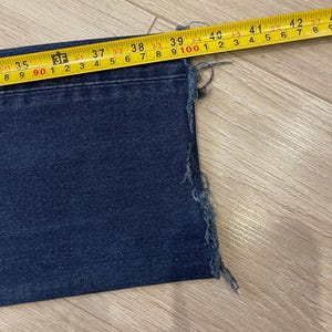 May include: Close-up of a section of dark blue denim jeans with a frayed edge. A yellow measuring tape shows the jeans are approximately 39 inches wide. The jeans are lying on a light brown wooden surface.