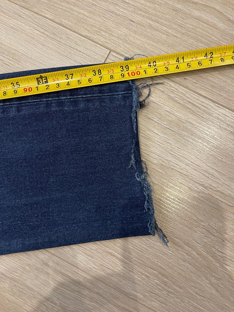 May include: Close-up of a section of dark blue denim jeans with a frayed edge. A yellow measuring tape shows the jeans are approximately 39 inches wide. The jeans are lying on a light brown wooden surface.