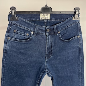 May include: Dark blue denim jeans hanging on a black hanger. The jeans have a classic five-pocket design with silver rivets and a button closure. The label reads "Acne Studios".