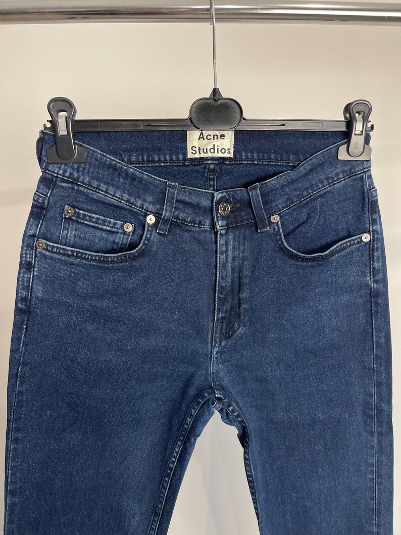 May include: Dark blue denim jeans hanging on a black hanger. The jeans have a classic five-pocket design with silver rivets and a button closure. The label reads "Acne Studios".