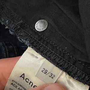 May include: Close-up of dark denim jeans with a silver button that reads "Acne Studios". A white tag displays the brand name "Acne" and size "29/32". The jeans are made of a dark wash denim material.