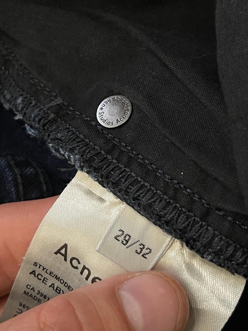 May include: Close-up of dark denim jeans with a silver button that reads "Acne Studios". A white tag displays the brand name "Acne" and size "29/32". The jeans are made of a dark wash denim material.