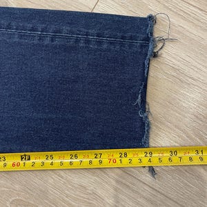 May include: Close-up of a section of dark blue denim jeans with a raw, frayed hem. A yellow measuring tape shows the jeans are approximately 29 inches wide at the hem.