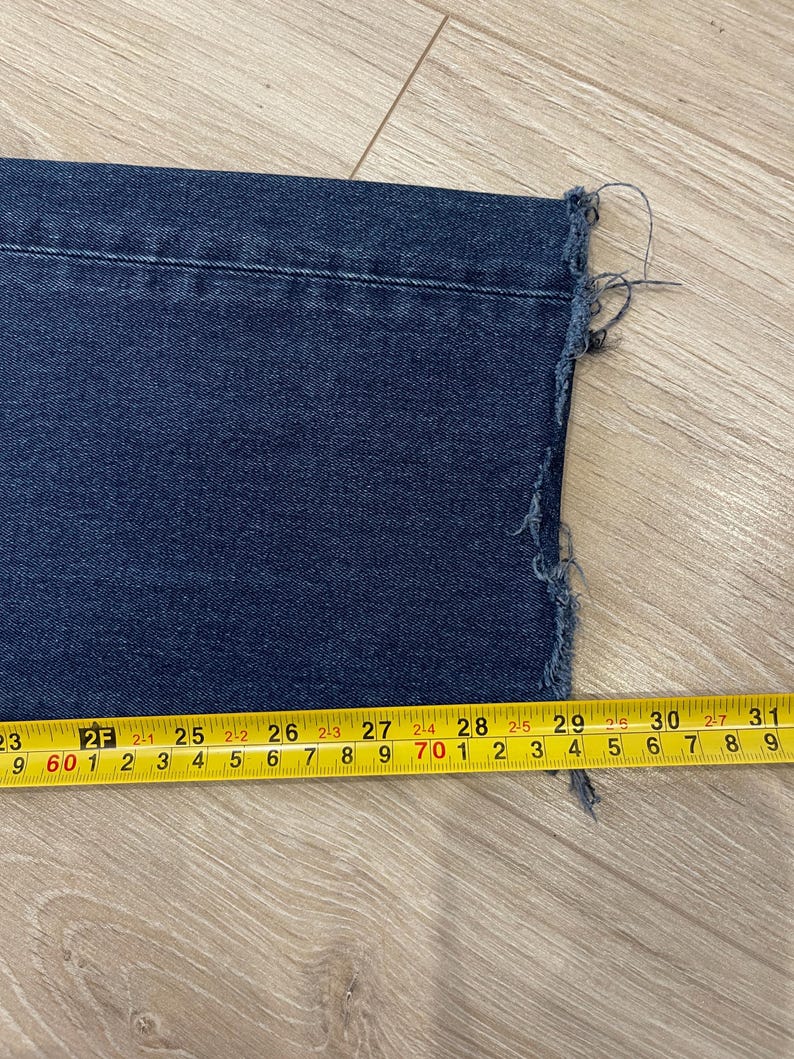 May include: Close-up of a section of dark blue denim jeans with a raw, frayed hem. A yellow measuring tape shows the jeans are approximately 29 inches wide at the hem.