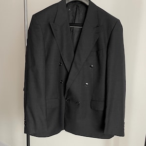 Può includere: Un blazer nero doppiopetto appeso a un attaccapanni. Il blazer ha un revers a lancia, due file di bottoni e due tasche anteriori. Il blazer è realizzato in tessuto scuro ed è uno stile classico.