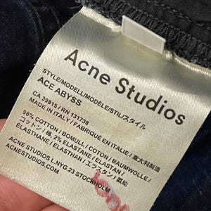 May include: Close-up of a white tag from Acne Studios, with the brand name and "ACE ABYSS" style details. The tag includes fabric composition, country of origin, and website address. The tag is attached to dark blue denim.
