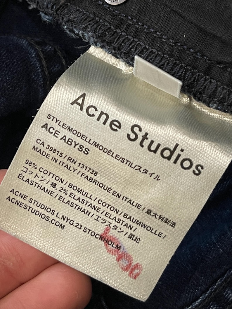 May include: Close-up of a white tag from Acne Studios, with the brand name and "ACE ABYSS" style details. The tag includes fabric composition, country of origin, and website address. The tag is attached to dark blue denim.
