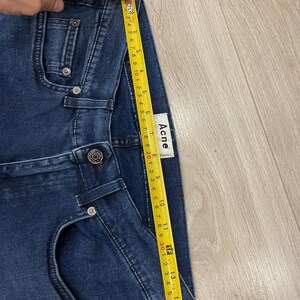 May include: Dark blue denim shorts with a yellow measuring tape showing a length of approximately 15 inches. The shorts have a button closure and pockets. The brand label "Acne" is visible.