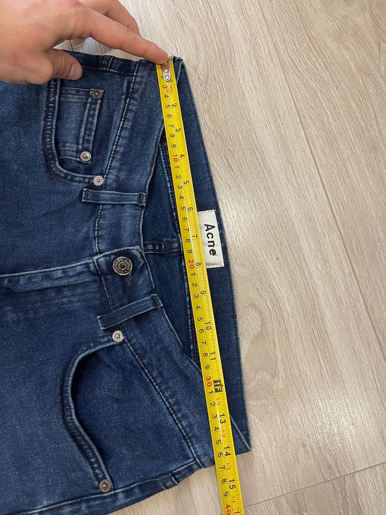 May include: Dark blue denim shorts with a yellow measuring tape showing a length of approximately 15 inches. The shorts have a button closure and pockets. The brand label "Acne" is visible.