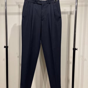 Może przedstawiać: Granatowe spodnie z zakładkami wiszące na wieszaku. Spodnie mają zapięcie na guziki, szlufki i przednie kieszenie. Mankiety są podwinięte u dołu. Spodnie wykonane są z gładkiej, jednolitej tkaniny.