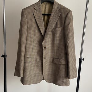 Può includere: Un blazer color marrone e beige con motivo pied-de-poule, caratterizzato da risvolti classici, due bottoni frontali e due tasche con patta. La giacca presenta una fodera interna beige ed è appesa a una gruccia nera. Il tessuto ha un motivo a quadri.