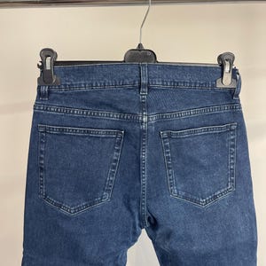May include: Dark blue denim jeans hanging on a hanger. The jeans have two back pockets and a classic five-pocket design. The denim appears to be a medium weight, with visible stitching details.