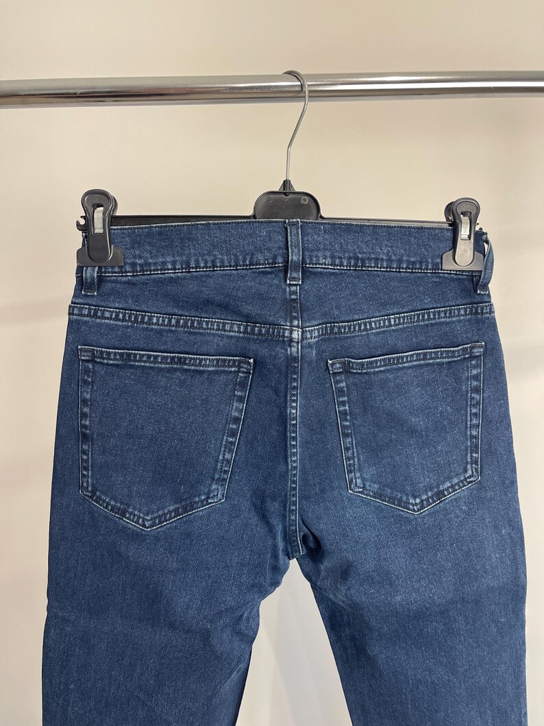 May include: Dark blue denim jeans hanging on a hanger. The jeans have two back pockets and a classic five-pocket design. The denim appears to be a medium weight, with visible stitching details.